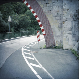 Eine bogenförmige Durchfahrt unter einer Eisenbahnbrücke ist mit rot-weißen Markern versehen. Gegen eine Straßenmarkierung bilden sich zwei harmonierende Bögen, die das Bild markant dominieren.