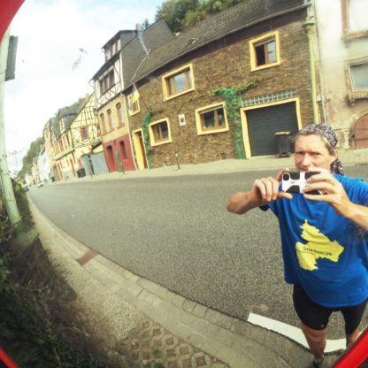 Handyfotografierender Mensch mit blauem T-Shirt spiegelt sich in einem innerörtlichen Straßenspiegel vor geschlossener Häuserzeile am Straßenrand. Auf dem T-Shirt ist ein gelber Umriss, der Silhouette des Bundeslands Rheinland-Pfalz, spiegelverkehrt.