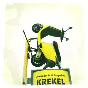 Gelber Motorroller, der senkrecht auf einem hoch in die Luft ragenden Firmenschild montiert ist und in den weißen Himmel ragt. Firmenname auf dem Schil lautet Krekel.