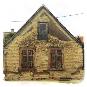Gibel eines alten Hauses mit einem großen Fenster mittig über zwei großen Fenstern. Der lehmfarbene Verputz blättert ab.