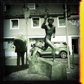 Schwarzweißbild eines Bronzedenkmals, rennender Junge vor innerstädtischer Fußgängerampel. Mit dem Rücken zu Jüngling und Betrachterin steht ein wuchtiger Mann mit Anzug an der Ampel. Ins Schwarzweiß brennt sich von rechts fehlfarbig ein gelborangener Belichtungsfehlerrand.