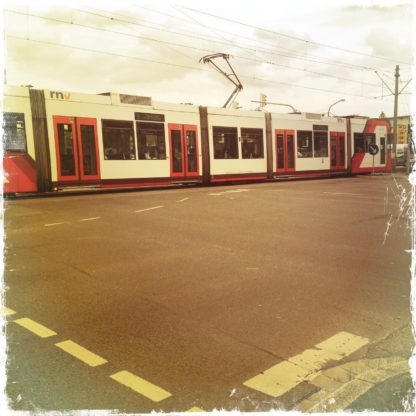 Im oberen Drittel des quadratischen Bilds durchquert eine rot-weiße Straßenbahn die Szene. Die beiden unteren Drittel zeigen Teer, Straße, Markierungen in einem rötlichen Teint.