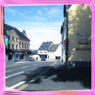 Innenstadtszene mit Straße links im Bild, halb angeschnitten. Rechte Bildseite eine Hausfassade mit Schriftzügen. Das bIld hat einen rosa Rahmen und ist blaugrau getönt. Ein nicht sichtbarer Baum rechts wirft einen großen Schatten auf den Teer.
