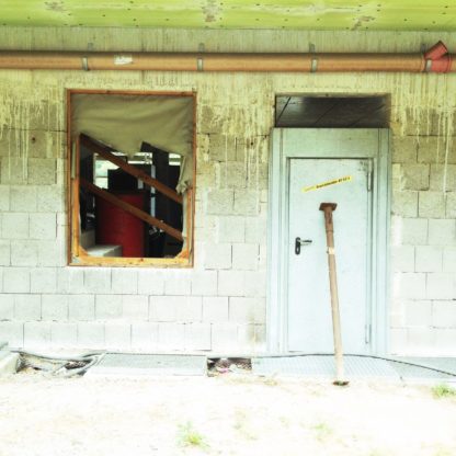 Baustelle mit frischer Fassade unter Vorbau. Oberhalb schattiert. Die Tür ist mit einer schräg stehenden Baustütze versperrt. Im spiegelnden Fenster links daneben stehen Gegenstände.