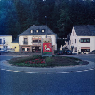 Kreisverkehr zentral im Bild vor Walm bedachtem Haus. Auf dem Kreisel ein rotes Stadtwappen mit weißem Motiv