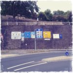 Viele rechteckige Hinweisschilder auf Autobahnen und Bundesstraßen stehen in Reih und Glied vor einer hohen Mauer, über der sich eine Art Streuobstwiese befindet.