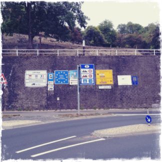 Viele rechteckige Hinweisschilder auf Autobahnen und Bundesstraßen stehen in Reih und Glied vor einer hohen Mauer, über der sich eine Art Streuobstwiese befindet.