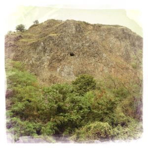 Winziger schwarzer Punkt in wuchtigem Schieferfelsen über wildem Baumbewuchs. Der Punkt ist der Eingang einer Höhle mitten in der Steilwand.