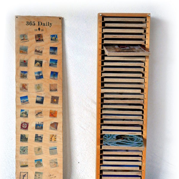 Offene Holzbox, Deckel steht links daneben. Aus den Fächern mit Kunstwerken ragen zwei etwa heraus wie Schubladen. Box Artists Collection.