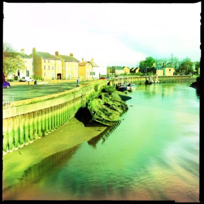 Blick auf einen Meernahen Flusshafen bei Ebe. Grünstichig fehlfarbig liegt ein alter Holzkahn auf Schlick an der Hafenmauer vertäut. Im Hintergrund englische Stadtszene.
