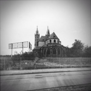 Scharzweiß-Bild eines Kirchbaus in einem flachen Rebenland neben einer Bundesstraße. Großes Domänenhinweisschild zwischen den Reben.