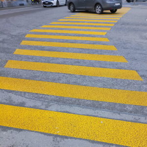 Bildfüllender gelber Zebrastreifen, der sich nach rechts knickt. Ganz oben Reifen und unterer Teil eines grauen und eines weißen PKWs, die die Straße queren.