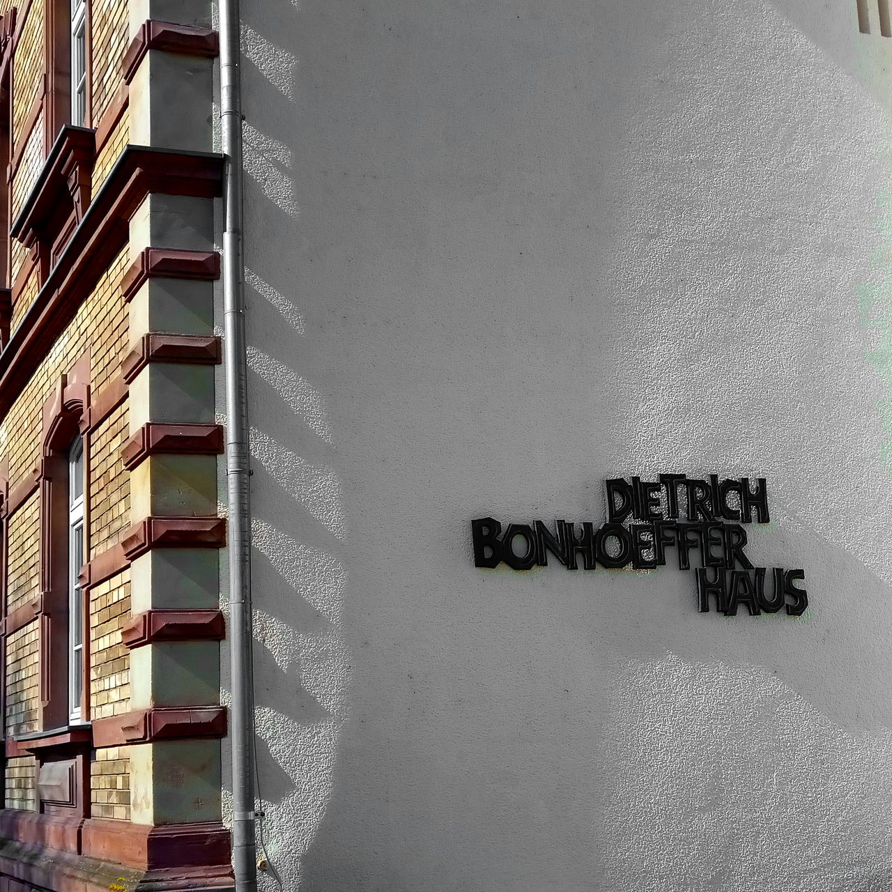 Die Aufschrift Dietrich Bonhoefer Haus aus gusseisernen schwarzen Lettern an der weißen Wand einer Hausecke, die günstig vom Licht und Schatten der gezackten Mauerecke und Dachrinne gestreift wird und somit eine unvermutete Tiefe erfährt.