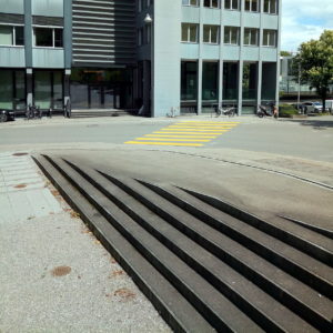Weitläufige Außentreppe im unteren Teil des Bildes läuft längst zu einem gelben Zebrastreifen vor einem Geschäftsgebäude. Ein paar Mopeds und Fahrräder stehen auf dem Gehweg