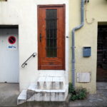 eine Steile, enge, weiß gekachelte, dreistufige Haustreppe führt zu einer braunen Tür, deren Kern verglast und vergittert ist. Daneben ein Regenfallrohr, ein Briefkasten und die Einfahrten zu Nachbargaragen.