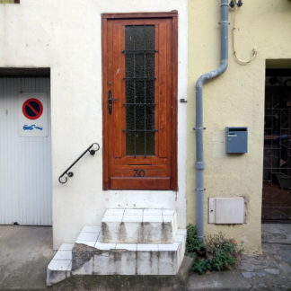 eine Steile, enge, weiß gekachelte, dreistufige Haustreppe führt zu einer braunen Tür, deren Kern verglast und vergittert ist. Daneben ein Regenfallrohr, ein Briefkasten und die Einfahrten zu Nachbargaragen.
