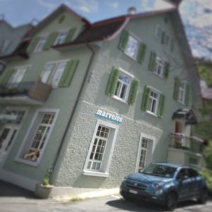 Ein typisches schweizerisches Stadthaus, das in unscharfer Vignette und schräg abgebildet einen Horror-Haus-Touch erhält. Der Schriftzug "marvelos" über einem Fenster im Erdgeschoss deutet an, dass sich ein Fahrradladen hinter den Mauern verbirgt. Vorm betreppten Hauseingang steht ein blaues Auto zeitgenössicher Bauart.