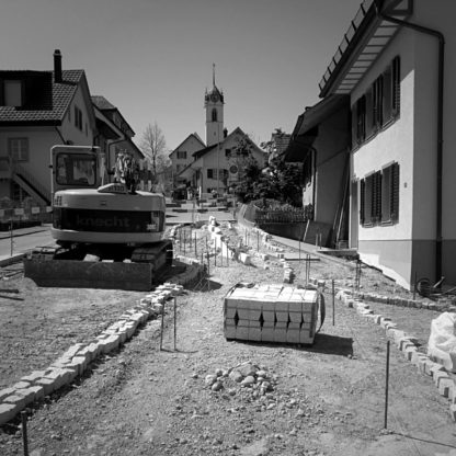 Aufgerissene Dorfstraße, die auf eine Kirche hinzu führt. Die Ränder der Straße sind mit Pflastersteinen provisorisch gelegt, Links ein Bagger. Viele Eisenstäber und Schnüre zeigen die Bauvermessung. Niemand arbeitet.