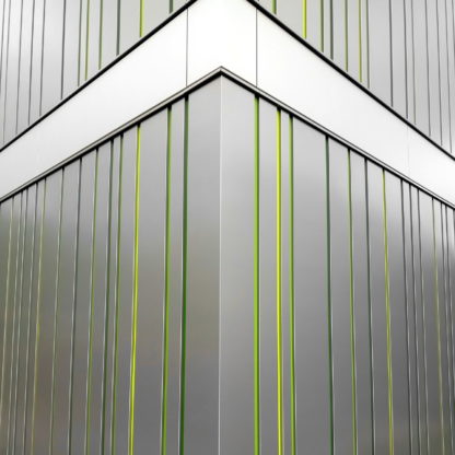 Moderne Fassade auf die Hausecke fokussiert. Graue metallisch glänzende Elemente werden senkrecht gebrochen von grünen Fugen. Eine horizontal verlaufende Fläche unterbricht die senkrechten Elemente und lässt die Szene pfeilförmig wirken.