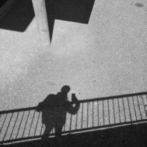 Der Schatten eines Menschen, der mit dem Handy fotografiert und an einem Geländer lehnt. Nur teer und Schatten und im Ansatz eine schmale runde Säule, die wiederum Schatten wirft.