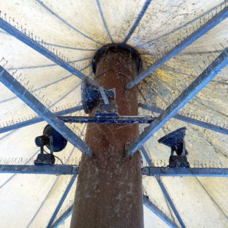 Blick von unten auf eine Stahlstütze, die mit eisernen Armen eine Art Schirm trägt. Auf den Querarmen befinden sich Stacheln, um Tauben zu vergrämen. Leuchten sind angebracht. Viele Spinnweben umgarnen die Konstruktion.