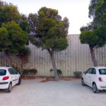 Zwischen zwei weißen Autos ist eine Lücke auf einem offenbaren Parkplatz vor einer lammelen-Metallwand. Pinien und deren Nadeln am Boden deuten auf einen mediterranen Ort hin.