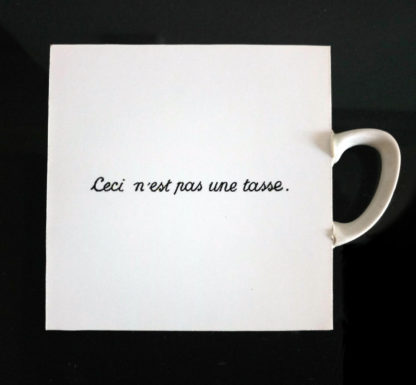 Quadratisches weißes Bild auf Holztafel mit der mittigen Aufschrift 'Ceci n'est pas une tasse'. Rechts klebt ein weißer Tassenhenkel an dem Objekt.