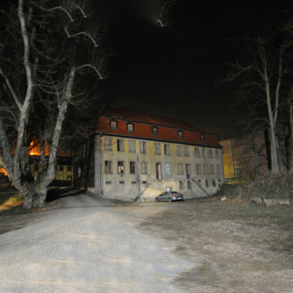 Nachtfotografie eines barocken Gebäudes neben kahlen Bäumen. Rotes Walmdach. Durch den Einsatz des Blitzgeräts zeichnet sich die Hoffläche vor dem Haus hell ab. Ein PKW parkt vor dem Eingang.