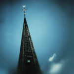 Die Spitze eines Kirchturms ragt bis fast zum oberen Bildrand in eine blaugrau düstere Athmosphäre. Der Hahn schimmert.