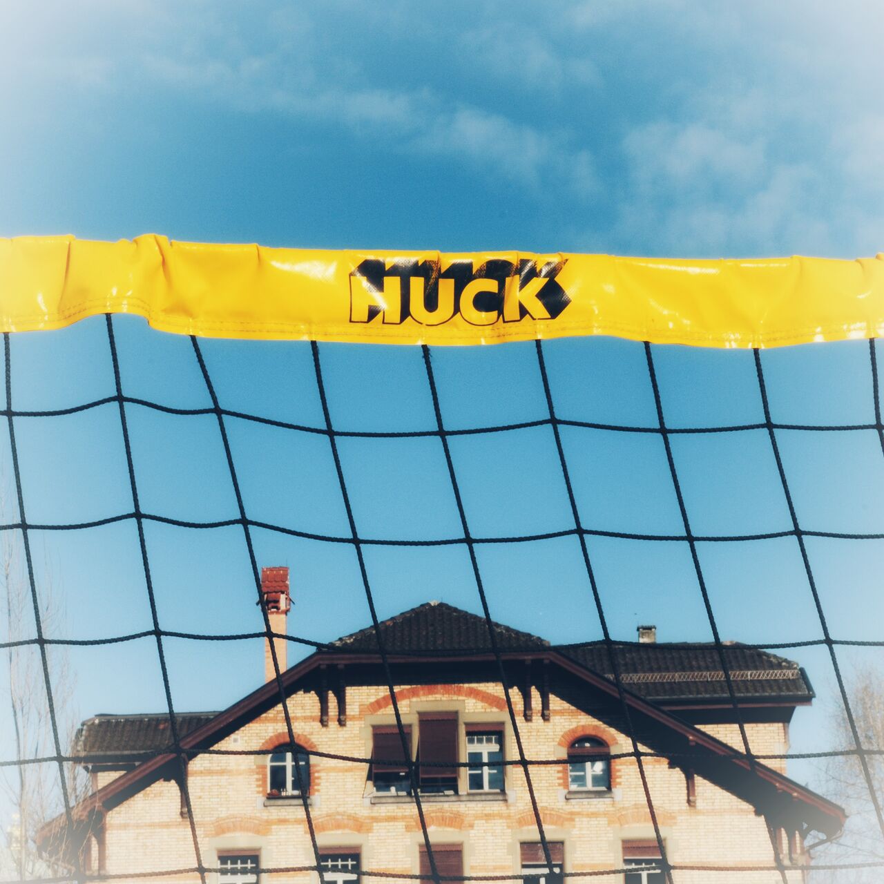 Blick durch ein Volleyballnetz mit gelber Kunststoffplanenkante auf einen Hausgiebel.