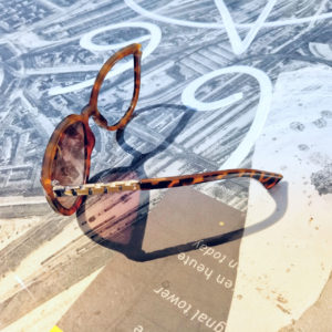 Brille liegt auf strukturierter Oberflche, die mit Schnörkeln in Blau- und Lavendeltönen versehen ist. Sonne wirft den Schatten nach rechts. Ein Bügel fehlt.