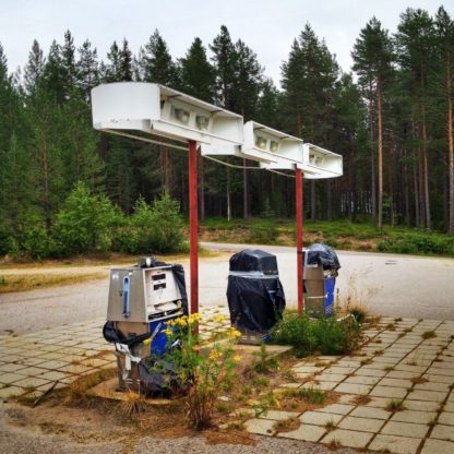 Unbedachte Tankstelle vor nordischem Wald, lange nicht mehr benutzt mit drei Zapfsäulen und kleinem, aber vandaliertem weißem Überbau