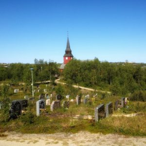 Über einen Friedhof zwischen Grabsteinen bahnt sich dein Blick den Weg durch geducktes Gesträuch, dem man die arktische Kargheit anmerkt. Am Horizont unter blauem Himmel ragt fern ein roter Kirchturm aus dem Grün