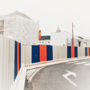 Rechtskurve innerstädtisch mit Richtungspfeil aufgemal. Die im Bau befindlichen Gebäude sind mit einer Wand aus weißen, blauen und orangenen Elementen abgetrennt
