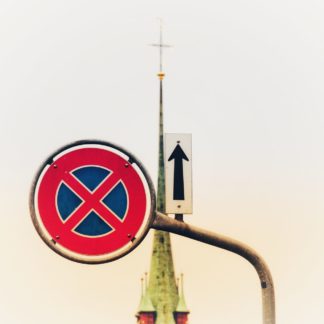 Zoomaufnahme eines Parkverbotsschilds mit einer Kirchturmspitze im Hintergrund. Ein Richtungspfeil, der nach oben zeigt, ergänzt das Ensemble.