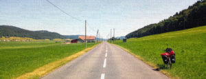 Fahrrad am rechten Straßenrand einer geraden Strecke in einem grünen Hochtal. Eine Überlandstromleitung begleitet die Straße. Weitab der Weiden sind bewaldete Berghänge.