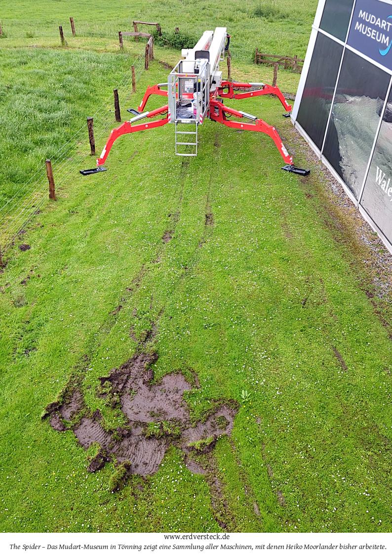 Neben einem Bauwerk steht auf grüner Wiese eine Hebebühne mit vier roten Füßen. Vor der Maschine befindet sich eine schlammige Spur, die das Gras signifikant verletzt. Blick von oben.